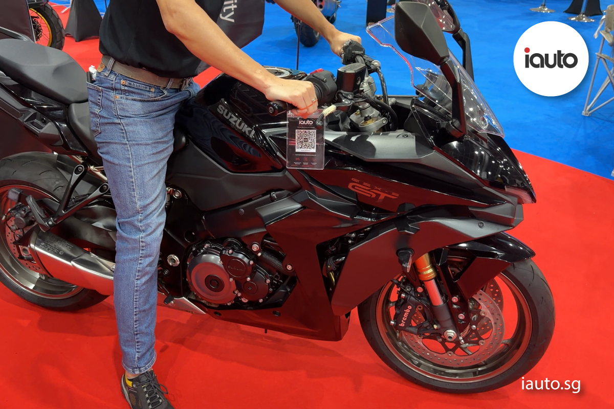 A person sitting on a black Suzuki GSX-8R motorcycle, which features a scannable iauto.sg QR code tag hanging from its right handlebar.