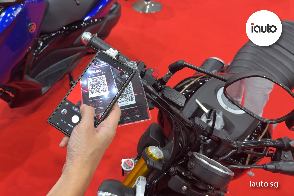A person holding a smartphone to scan a clear acrylic QR code display panel attached to the handlebars of a motorcycle, with the screen showing the iAuto digital platform.