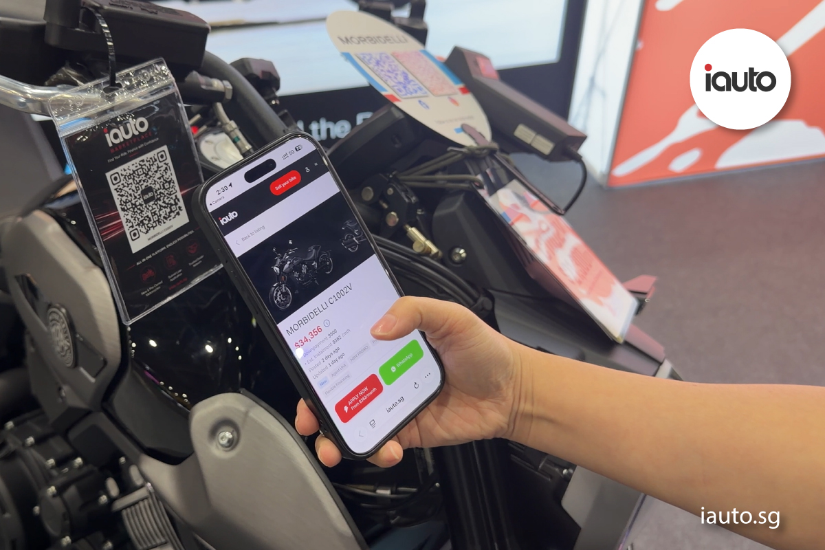 A person holding a smartphone to scan a clear acrylic iauto.sg QR code to browse vehicle details in iAuto Marketplace digital platform.