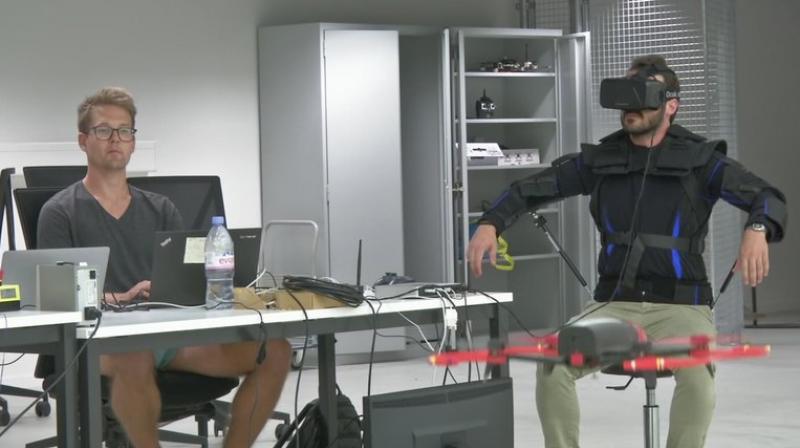 Robotics expert on the Ecole Polytechnique Federal de Lausanne, EPFL team, Matteo Macchini, right, displays the fly controls in a virtual reality scenario as he controls the flight of a drone in foreground, while observed by a fellow member of the team, July 23, 2018, in Lausanne, Switzerland. Scientists, engineers and robotics experts at the Swiss university have teamed up to develop a jacket that allows hands-free, torso-controlled flying of drones. (Ecole Polytechnique Federale De Lausanne via AP) MANDATORY DREDIT Robotics expert on the Ecole Polytechnique Federal de Lausanne, EPFL team, Matteo Macchini, right, displays the fly controls in a virtual reality scenario as he controls the flight of a drone in foreground, while observed by a fellow member of the team, July 23, 2018, in Lausanne, Switzerland. Scientists, engineers and robotics experts at the Swiss university have teamed up to develop a jacket that allows hands-free, torso-controlled flying of drones. (Ecole Polytechnique Federale De Lausanne via AP) MANDATORY DREDIT