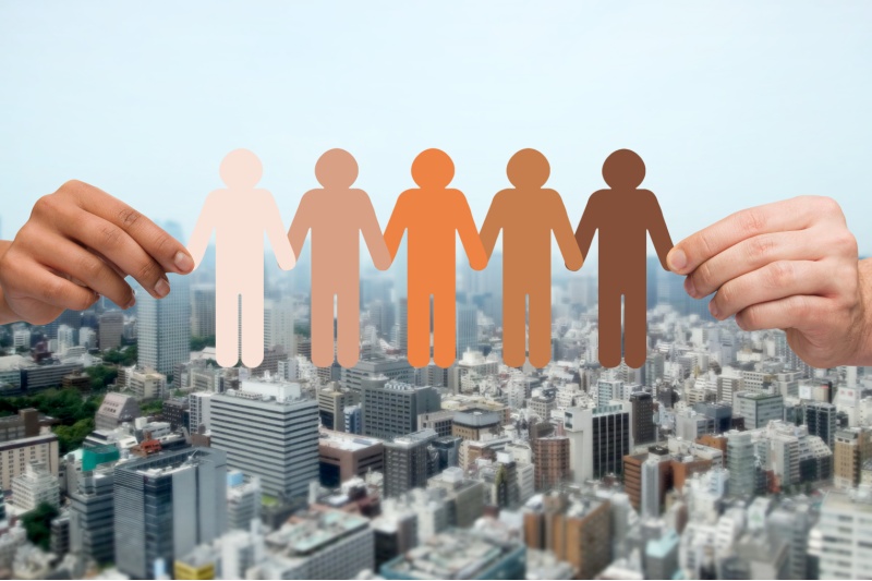 community, unity, population, race and humanity concept - multiracial couple hands holding chain of paper people pictogram over city background
