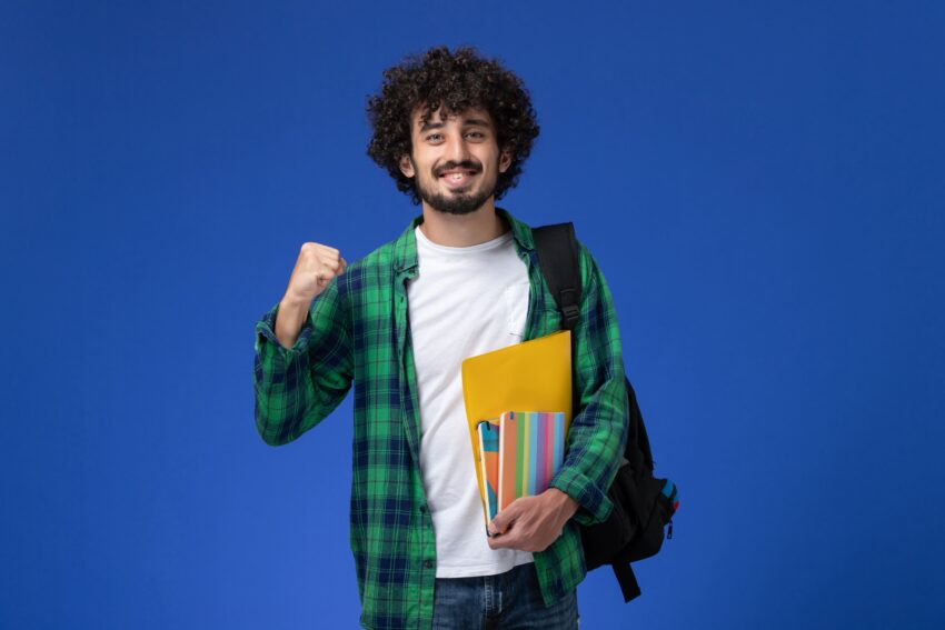 front view male student wearing black backpack holding copybooks files blue wall 1