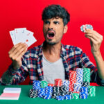 young hispanic man playing poker holding cards casino chips angry mad screaming frustrated furious shouting with anger rage aggressive concept