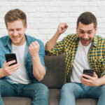 smiling young male friends looking smart phone clenching their fist