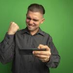 young handsome man grey shirt using gadget clenching fist happy excited rejoicing his success standing green wall