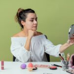 pleased beautiful girl sits table with makeup tools puts hand chin holding looking mirror isolated green wall 1