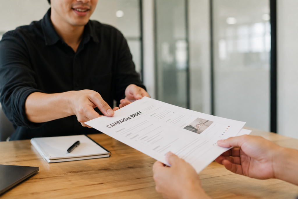 Person handing a printed campaign brief across a table during a project handoff meeting.