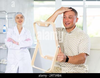nurse holds a mirror satisfied man examines his face in mirror after plastic surgery 2rt5rb4