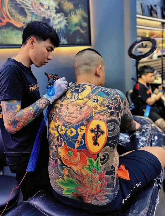 hình xăm kín lưng mèo thần tài samurai cho anh khách Người thợ xăm đang xăm hình chi tiết và sặc sỡ trên lưng khách hàng trong tiệm xăm.