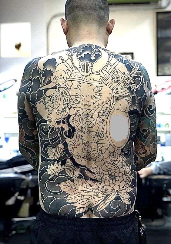 hoàn thiện hình xăm mèo thần tài samurai đơn sắc phủ kín lưng Người đàn ông xăm kín lưng với hình ảnh mèo thần tài và hoa văn phức tạp, đứng trong căn phòng sáng.