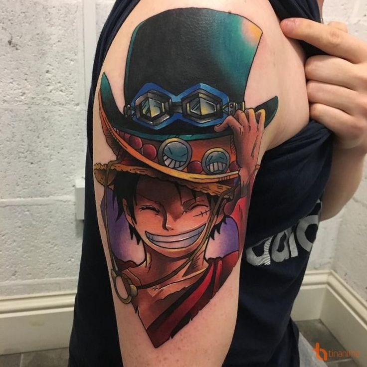 luffy với chiếc mũ thật cool Hình xăm trên cánh tay với nhân vật hoạt hình, đội mũ và cười tươi, nền tường xám phía sau.