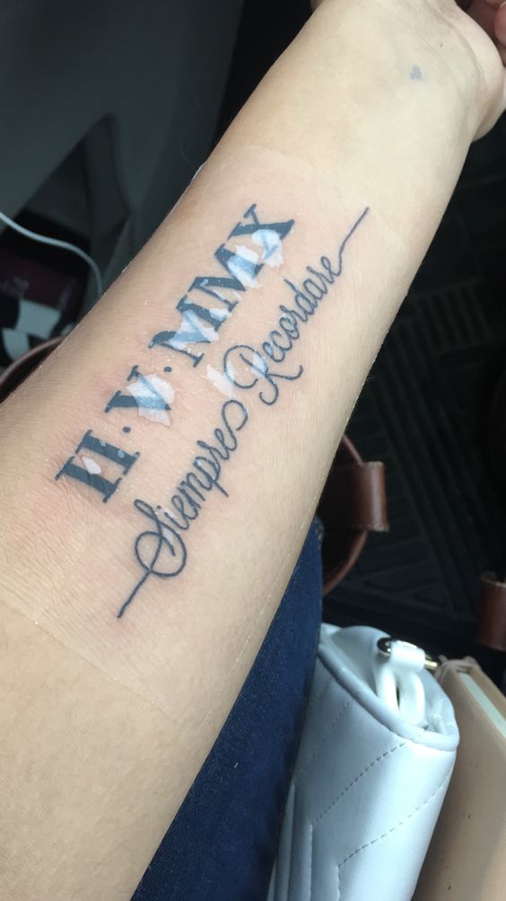 Hình xăm trên cánh tay với chữ số La Mã và dòng chữ viết tay, phía sau là túi xách màu trắng.