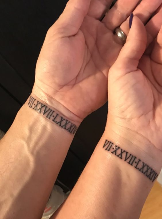 Hai cổ tay với hình xăm chữ số La Mã đen, hiển thị ngày tháng.