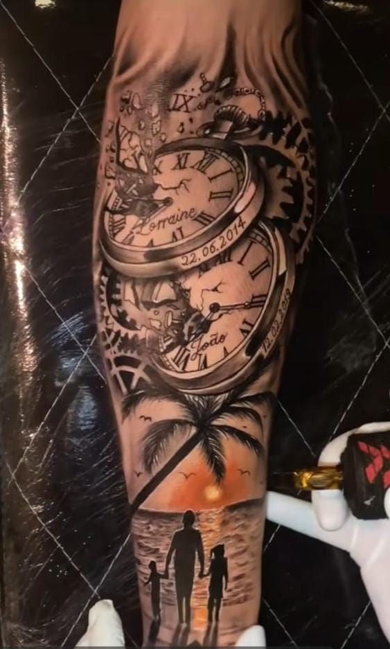 Hình xăm trên cánh tay với đồng hồ, bánh răng và cảnh hoàng hôn, phía dưới là bóng dáng gia đình trên bãi biển.
