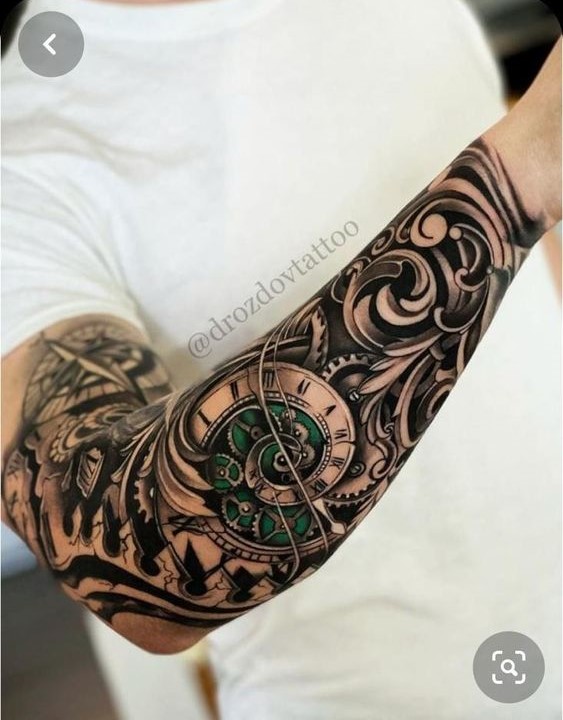 Cánh tay có hình xăm hoa văn phức tạp với hình đồng hồ và các chi tiết cơ khí màu xanh lá.