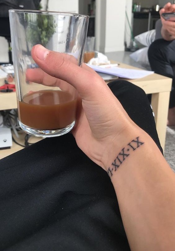 Một tay cầm ly thủy tinh chứa chất lỏng màu nâu, trên cổ tay có hình xăm số La Mã.
