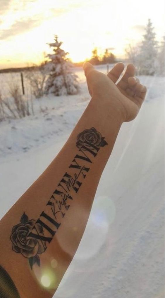 Cánh tay với hình xăm hoa hồng và số La Mã, nền là cảnh tuyết trắng và cây cối dưới ánh sáng mặt trời.