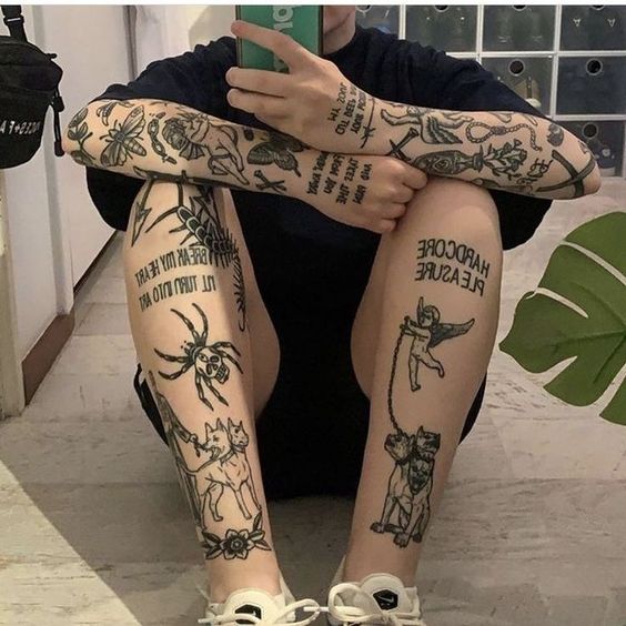 2c29a0885986bad2b705dae7794ab84d Người ngồi trên sàn, tay cầm điện thoại, trên cơ thể có nhiều hình xăm đa dạng, mặc áo tối màu và giày trắng.