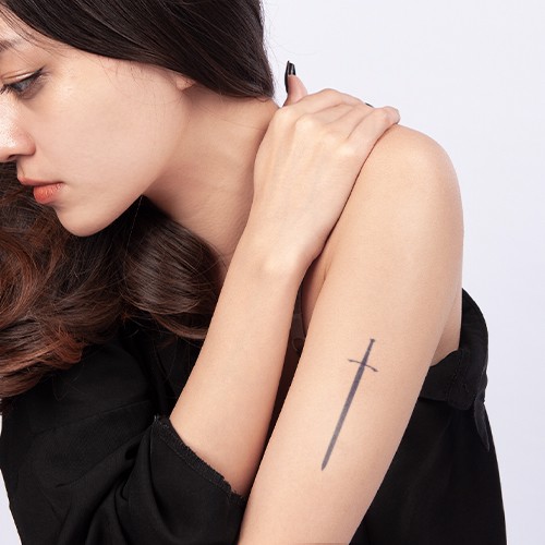 Người phụ nữ ngồi nghi&ecirc;ng, để lộ h&igrave;nh xăm thanh kiếm tr&ecirc;n c&aacute;nh tay, mặc &aacute;o đen, nền trắng.