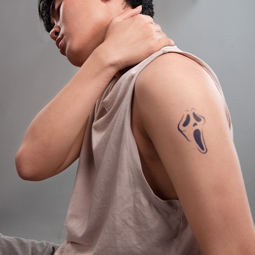 Người đang xoa cổ, tr&ecirc;n c&aacute;nh tay c&oacute; h&igrave;nh xăm mặt nạ kinh dị, mặc &aacute;o kh&ocirc;ng tay m&agrave;u s&aacute;ng.