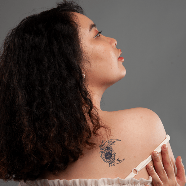 Người phụ nữ với m&aacute;i t&oacute;c xoăn, nh&igrave;n nghi&ecirc;ng, c&oacute; h&igrave;nh xăm hoa v&agrave; trăng lưỡi liềm tr&ecirc;n vai, nền x&aacute;m.