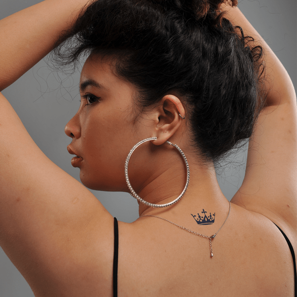 Người quay lưng, tay giơ l&ecirc;n đầu, đeo hoa tai lớn v&agrave; c&oacute; h&igrave;nh xăm vương miện tr&ecirc;n g&aacute;y.