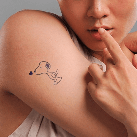 Người đang chỉ vào hình xăm nhỏ hình chú chó và trái tim trên cánh tay, mặc áo không tay màu trắng.