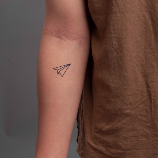 Cánh tay với hình xăm máy bay giấy nhỏ, áo màu nâu, nền xám.