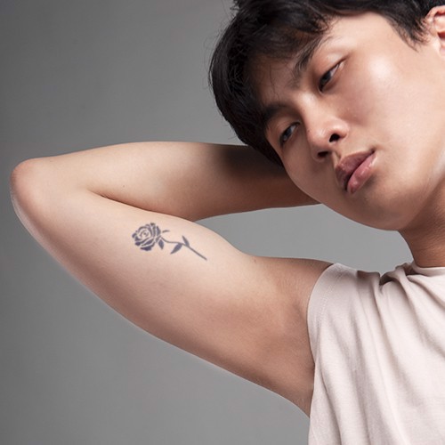 Người đang giơ tay lên đầu, trên bắp tay có hình xăm hoa hồng, nền xám nhạt.