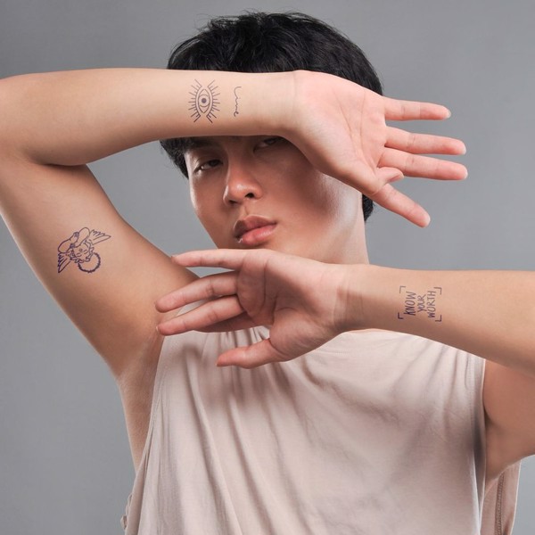 Người mặc áo sáng màu, hai tay đan chéo trước mặt, có hình xăm ở cánh tay và cổ tay.