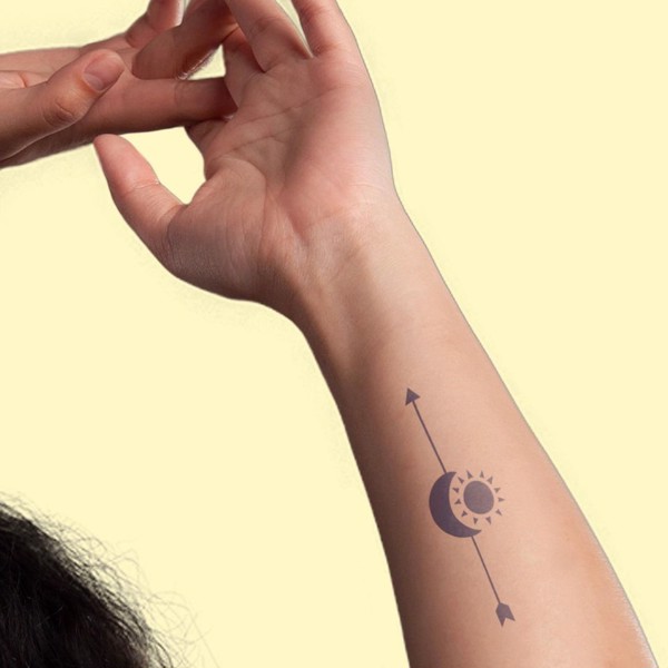 Cánh tay với hình xăm mũi tên và mặt trăng, mặt trời trên nền vàng nhạt.