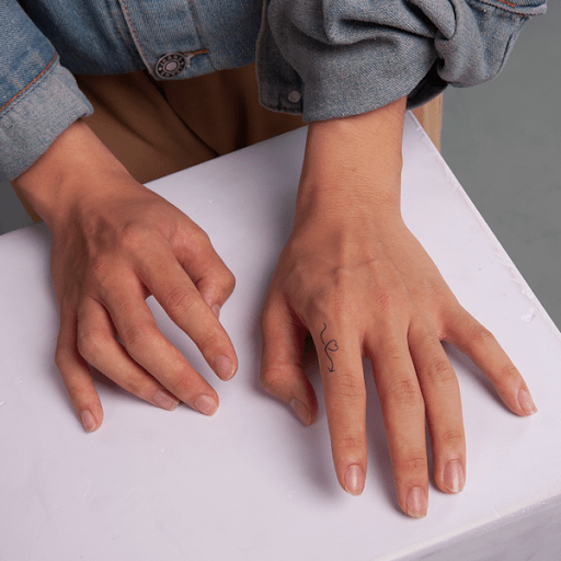 Hai bàn tay trên mặt bàn trắng, một tay có hình xăm nhỏ trên ngón áp út, mặc áo khoác jeans.