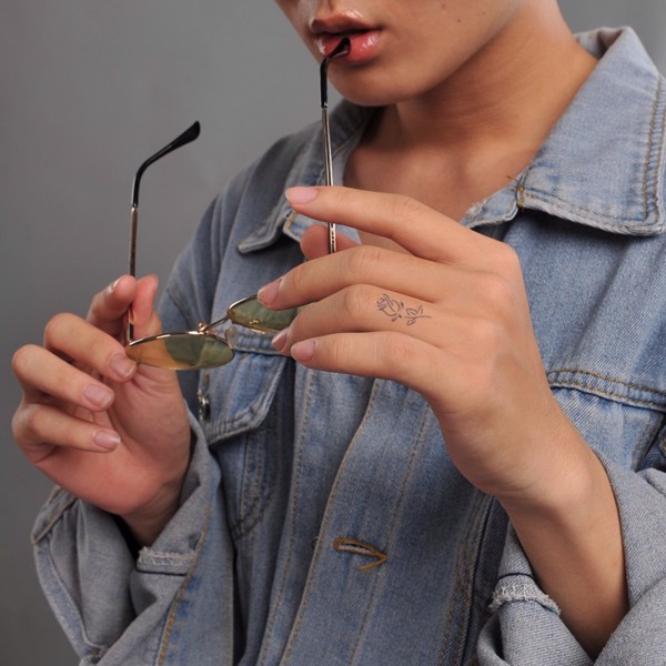 Người mặc áo khoác denim cầm kính râm, môi chạm gọng kính, tay có hình xăm nhỏ.
