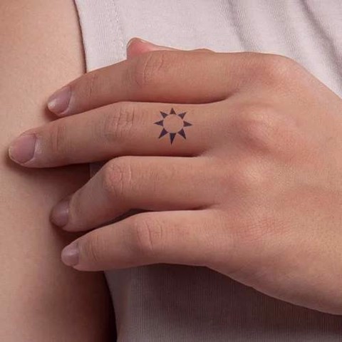 Bàn tay đặt lên vai, có hình xăm mặt trời nhỏ trên ngón tay giữa, nền áo màu nhạt.