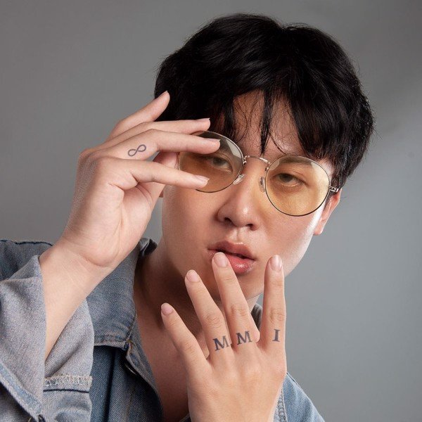 Người đeo kính râm, tóc đen, mặc áo denim, giơ tay có hình xăm ký hiệu và chữ số La Mã.