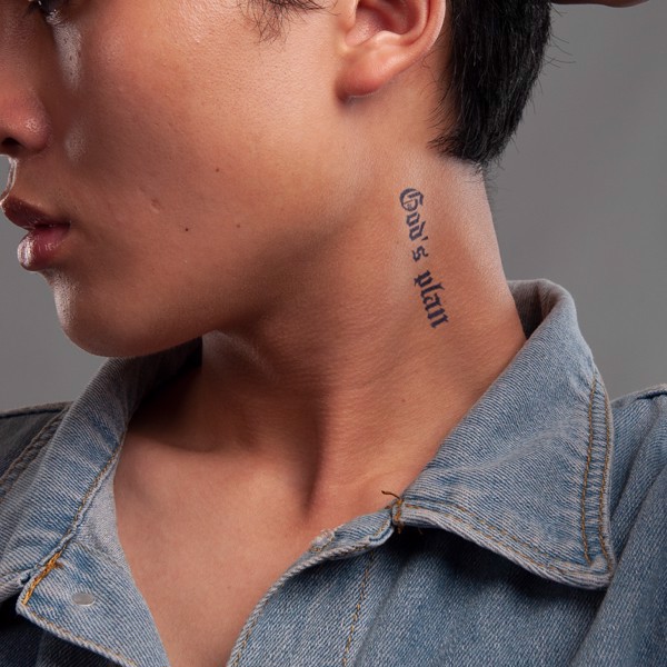 Cận cảnh một người đeo áo khoác denim với hình xăm chữ "God's plan" trên cổ.