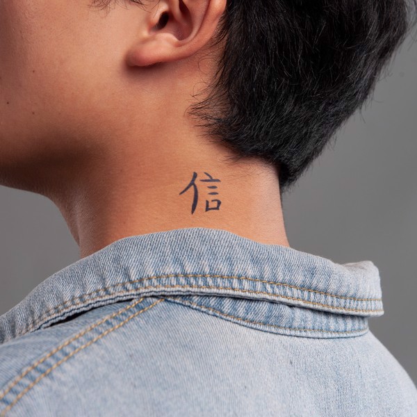 Người mặc áo denim có hình xăm chữ Hán nhỏ trên cổ, nền xám.