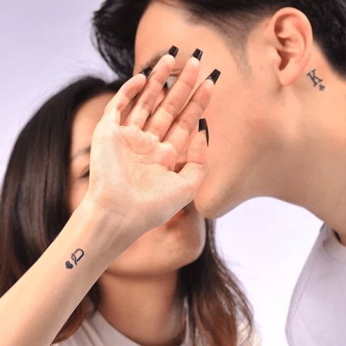 Người giơ tay với hình xăm quân bài, người khác có hình xăm tương tự ở cổ, cả hai đang tương tác gần gũi.