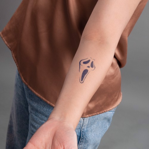 Cánh tay người có hình xăm mặt nạ kinh dị trên nền áo nâu và quần jeans.