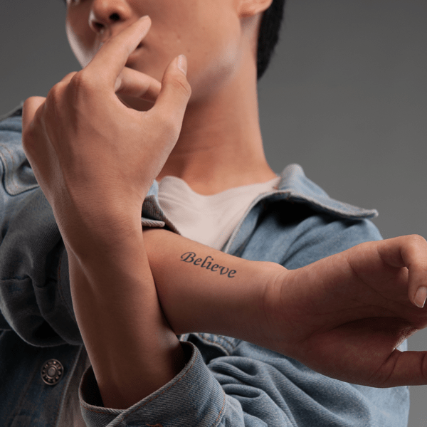 Người mặc áo khoác jean, tay có hình xăm chữ "Believe", đang giơ tay lên gần mặt trong khung cảnh trung tính.