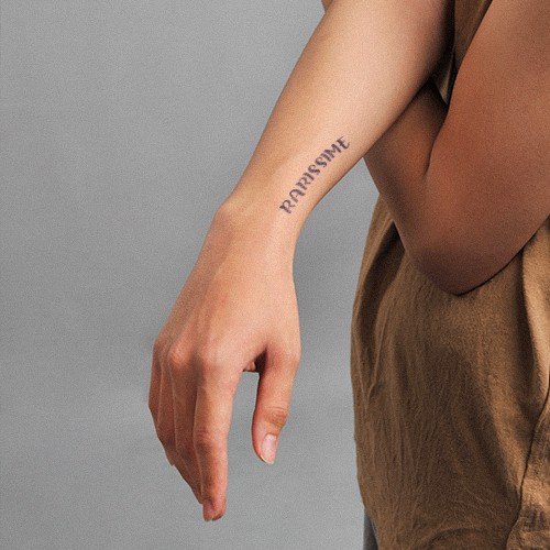 Cánh tay đeo áo nâu, có hình xăm chữ ở mặt trong cẳng tay, nền xám.