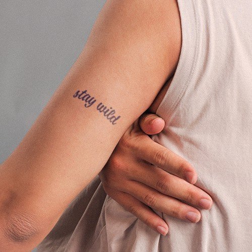 Cánh tay với hình xăm chữ "stay wild", người mặc áo không tay màu sáng.