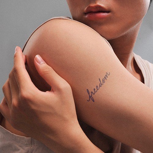 Người cầm cánh tay có hình xăm chữ "freedom" trên bắp tay, nền xám.