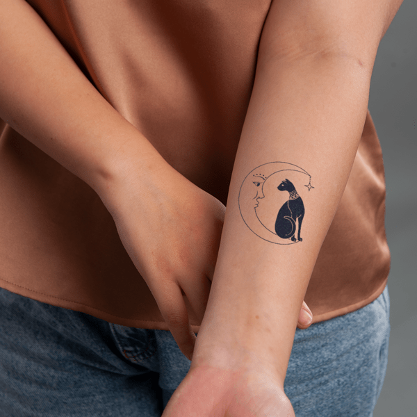 Hình xăm trên cánh tay, có thiết kế mặt trăng và mèo, được chụp trong phòng với nền xám.