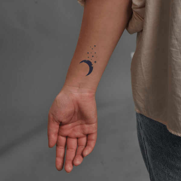 Cánh tay người với hình xăm mặt trăng và ngôi sao nhỏ trên nền xám.