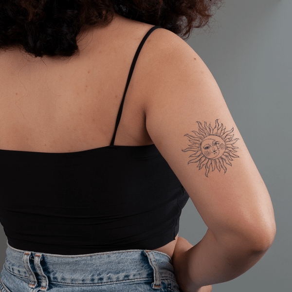 Người mặc áo đen, quần jeans, có hình xăm mặt trời trên cánh tay, nền màu xám.