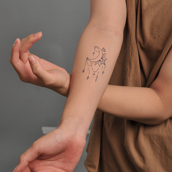 Hình xăm trên cánh tay với họa tiết mặt trăng và các ngôi sao, nền xám, áo màu nâu nhạt.