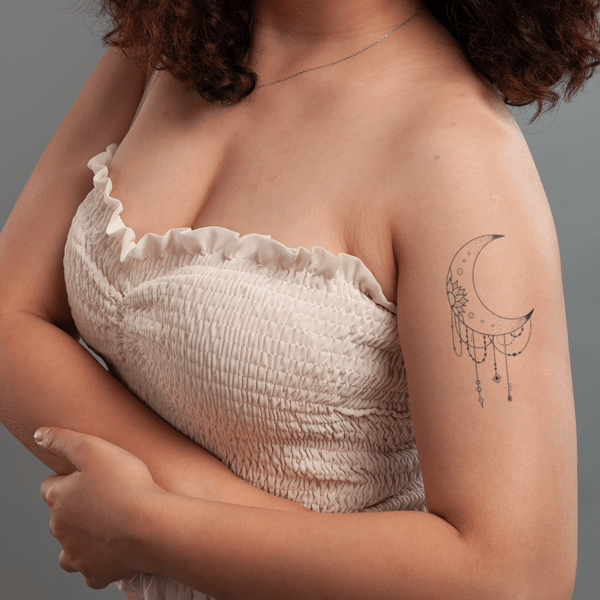 Người mặc áo trễ vai màu trắng, có hình xăm mặt trăng trên cánh tay, nền xám.