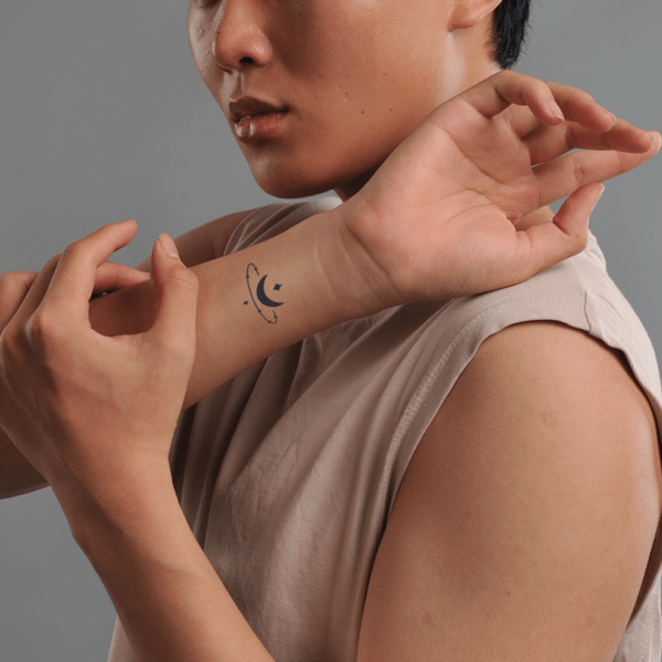 Người trong hình giơ cổ tay, có hình xăm nhỏ với họa tiết trăng và các vòng tròn xung quanh.