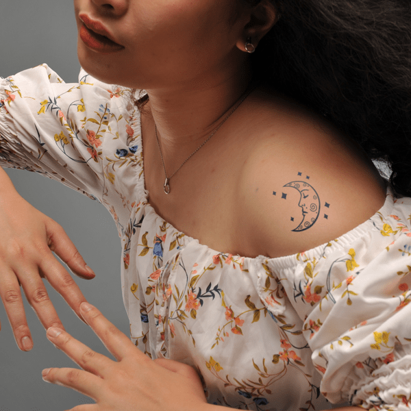 Người mặc áo hoa, có hình xăm mặt trăng trên vai trái, tay đặt nhẹ trên đùi.
